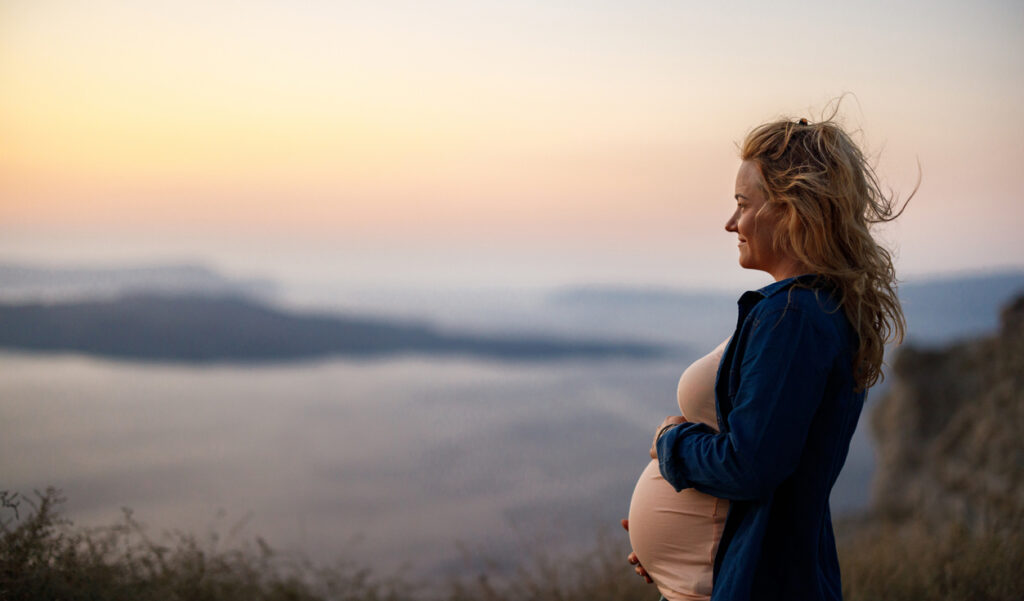 Smiling pregnant woman enjoying on a hill above the sea at sunset.