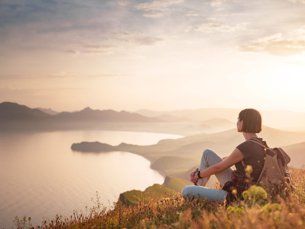 Woman sitting on a mountain top at sunset, symbolizing self-reflection, healing, and the journey of recovery.