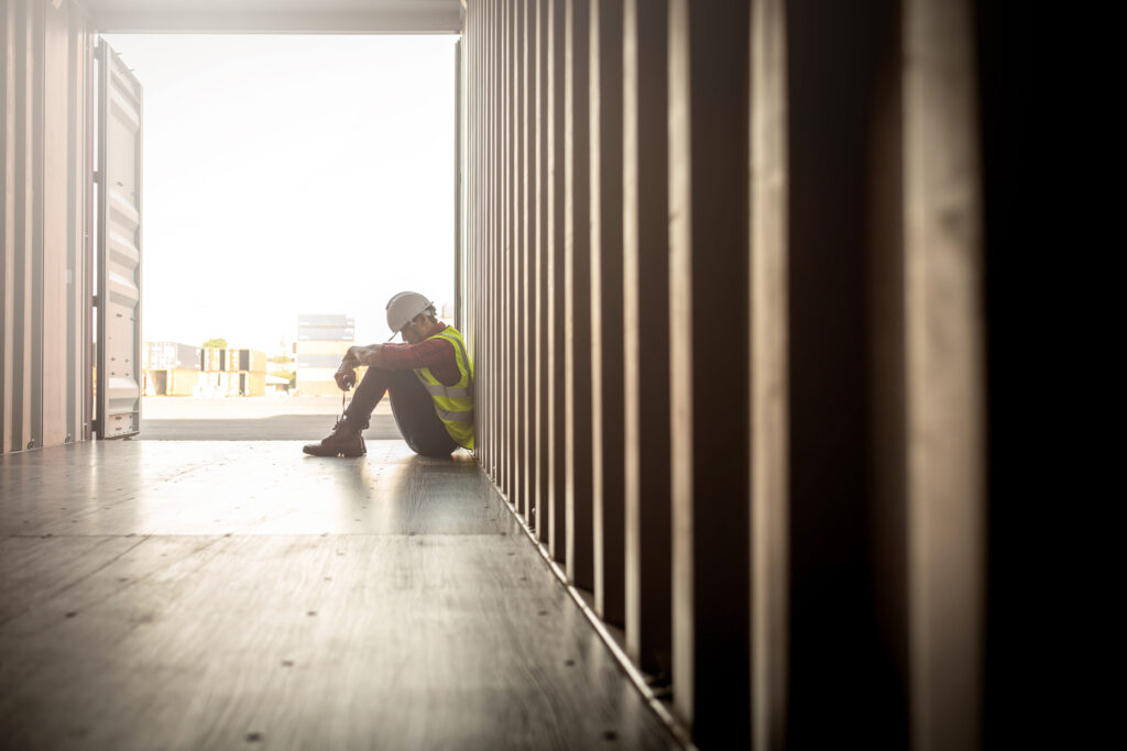 A construction worker sitting on a construction site, looking thoughtful or distressed, with safety gear in the background.