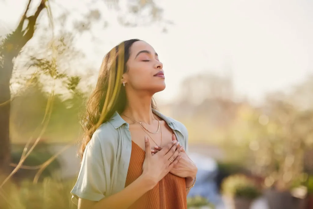 Young woman wearing a headband and standing outside with her hands on her heart, eyes closed feeling relaxed in nature