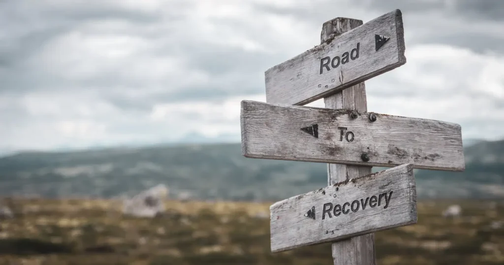 A wooden sign with three slats that together read "Road To Recovery" with nature in the background